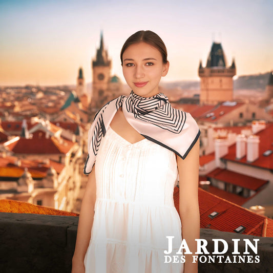Woman wearing a white dress with a black and white scarf in front of a cityscape, with 'Jardin des Fontaines' branding.